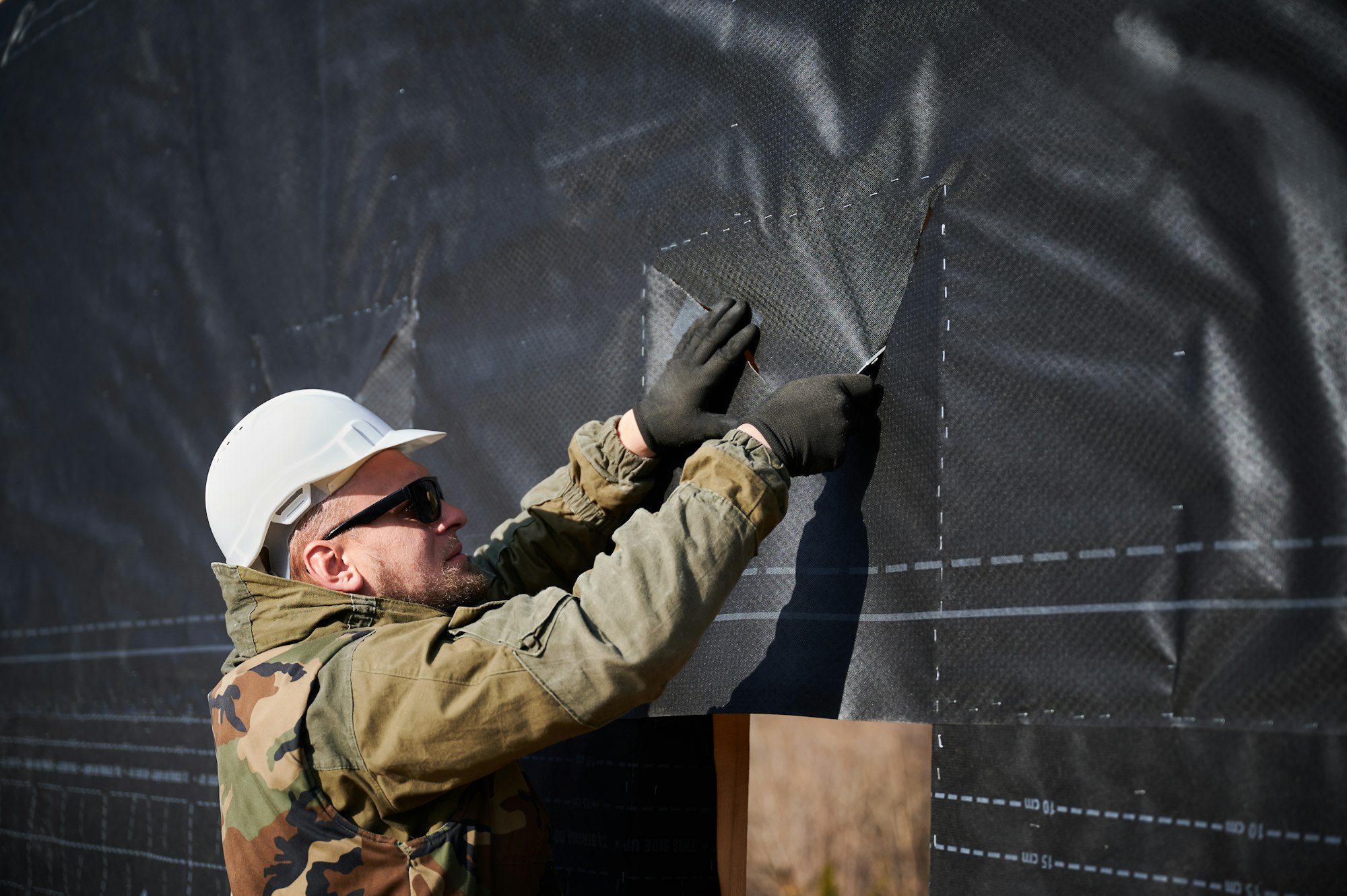 male builder installing waterproof membrane on wall of future wooden frame house